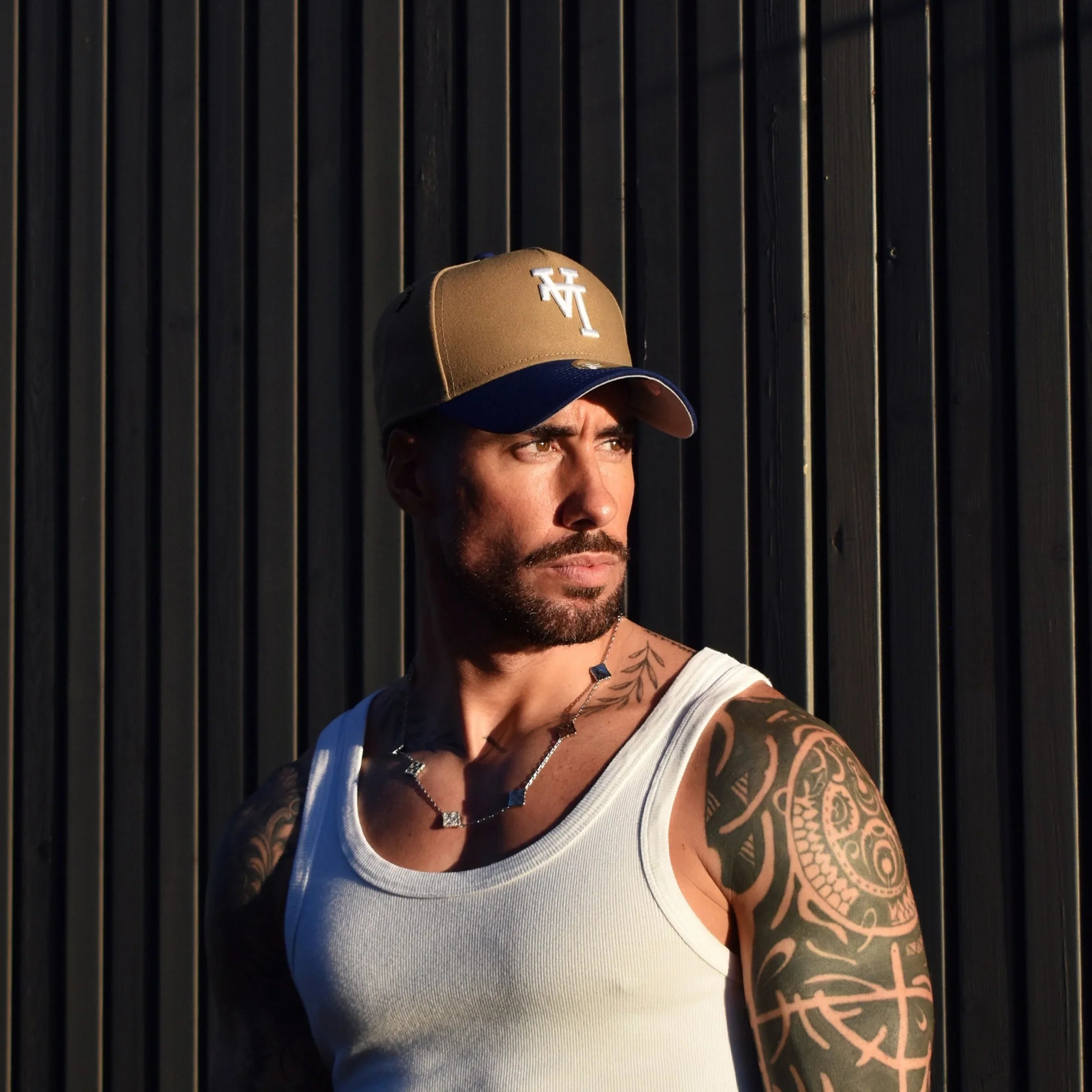 Man wearing a cap and tank top with tattoos on arms against a dark background