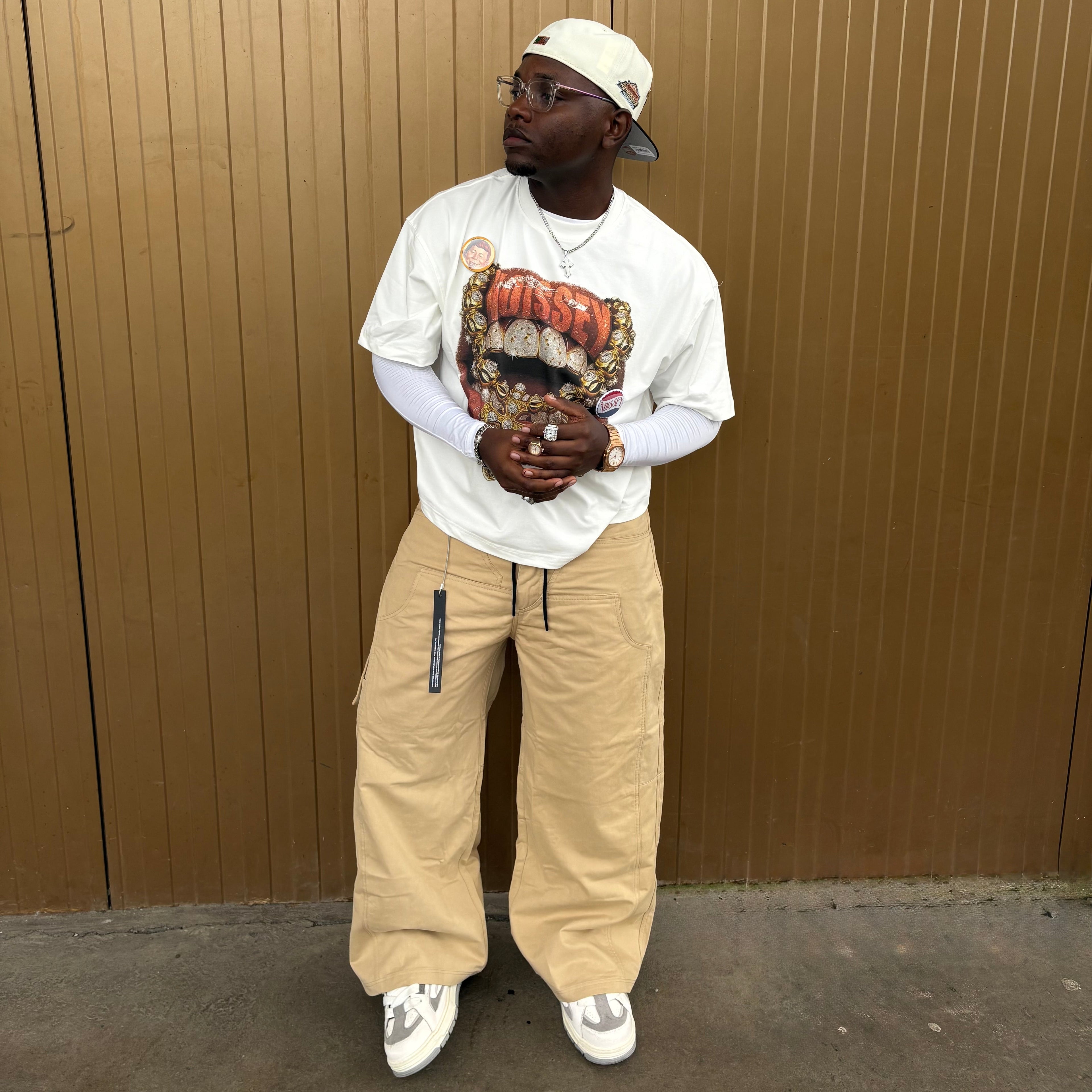 Person wearing a white t-shirt with graphic design, beige pants, and a cap standing against a brown wall.