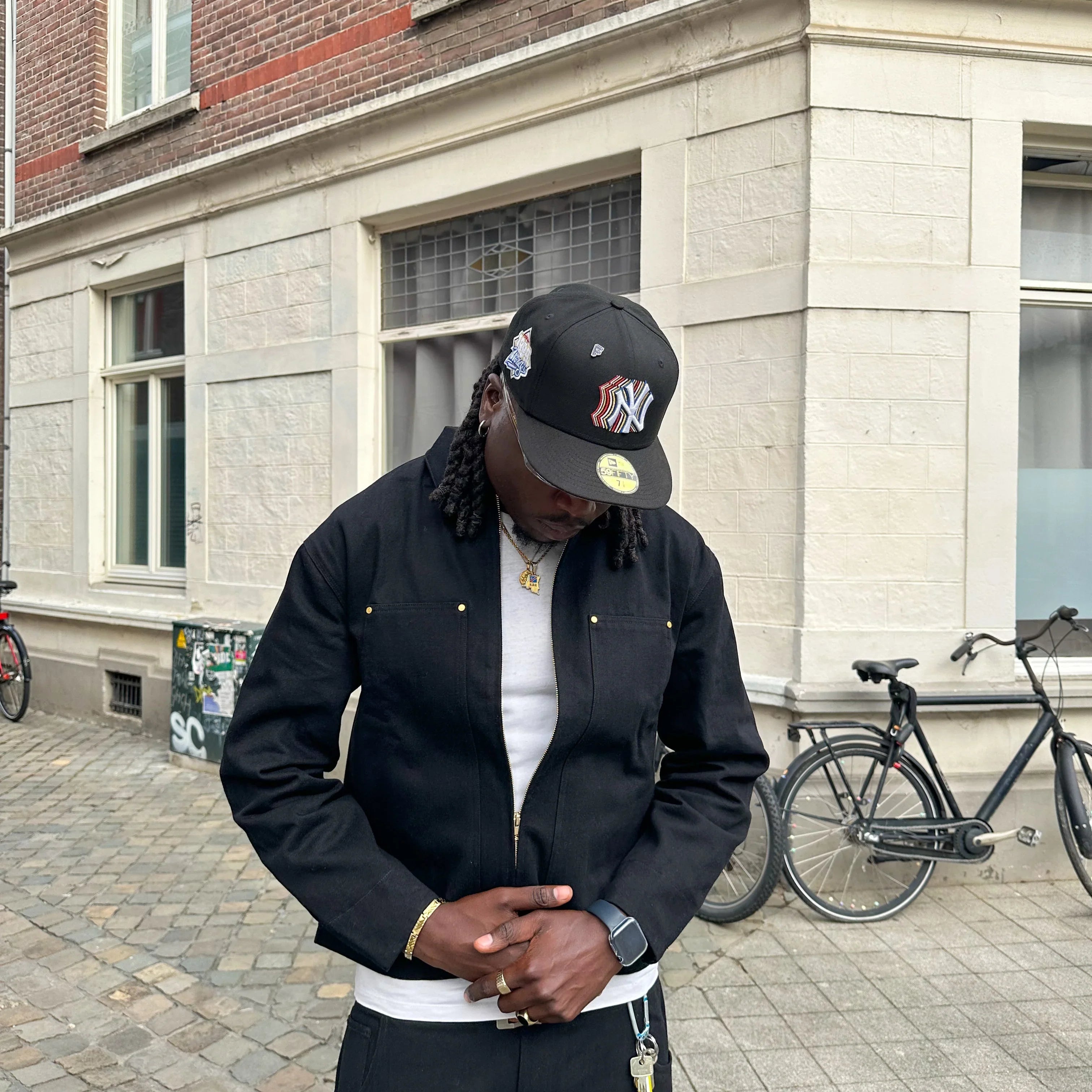 Person wearing a black jacket and cap in front of a brick building
