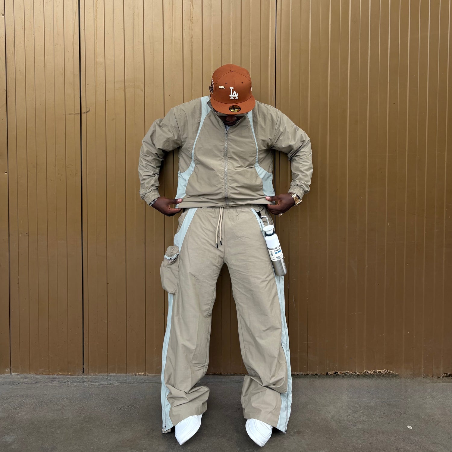 Person wearing a beige jumpsuit with white accents and a brown cap against a brown wall.