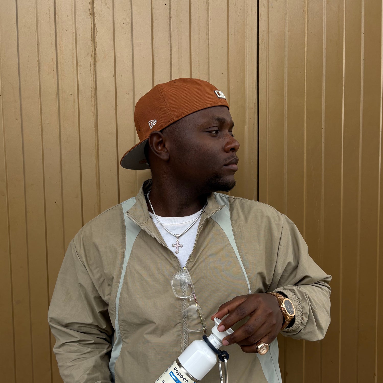 Man wearing a brown cap and beige jacket holding a white bottle against a wooden wall.