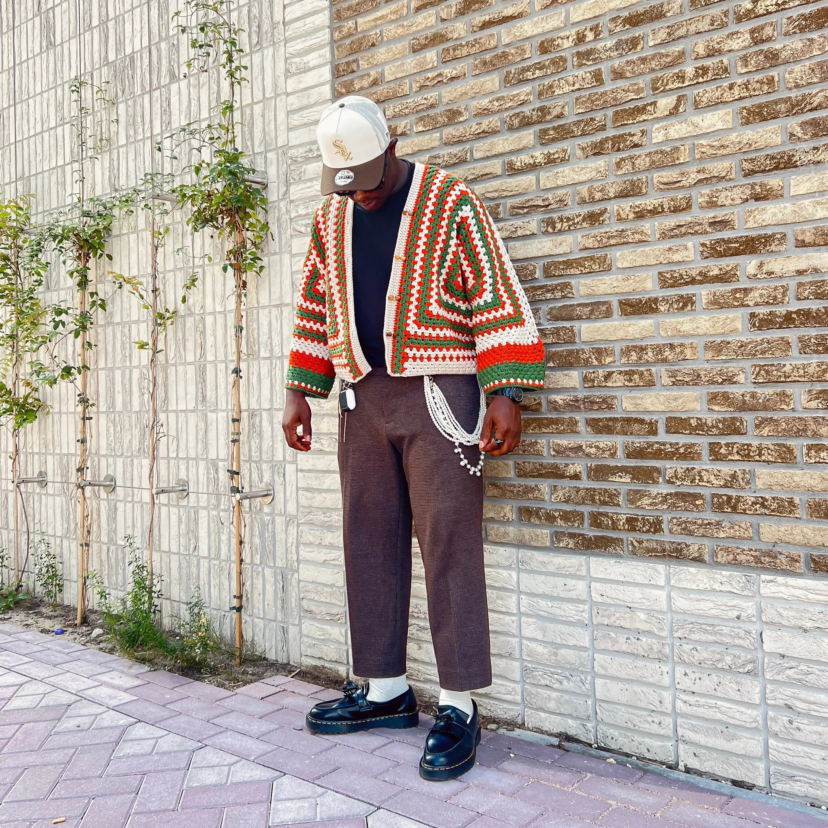 Person wearing a colorful knit cardigan, brown pants, and black shoes against a brick wall.